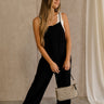 Full body front view of female model wearing the Jessie Wide Leg Jumpsuit in Black, that features black woven material, adjustable straps, and wide legs. Worn over white tank top.