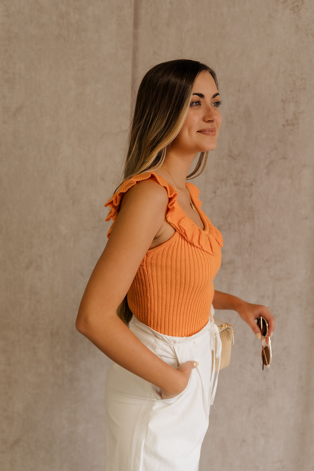 Side view of female model wearing the Lillian Orange Ruffle Tank which featuresOrange Knit Fabric, Scooped Neckline, Ruffle Details and Sleeveless.