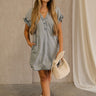 Full body view of female model wearing the Leah Denim Wash Tencel Mini Dress which features denim blue tencel fabric, mini length, two side slit pockets, a round neckline with a v-cutout, an elastic band in the back, and short sleeves with elastic hems and ruffle details.
