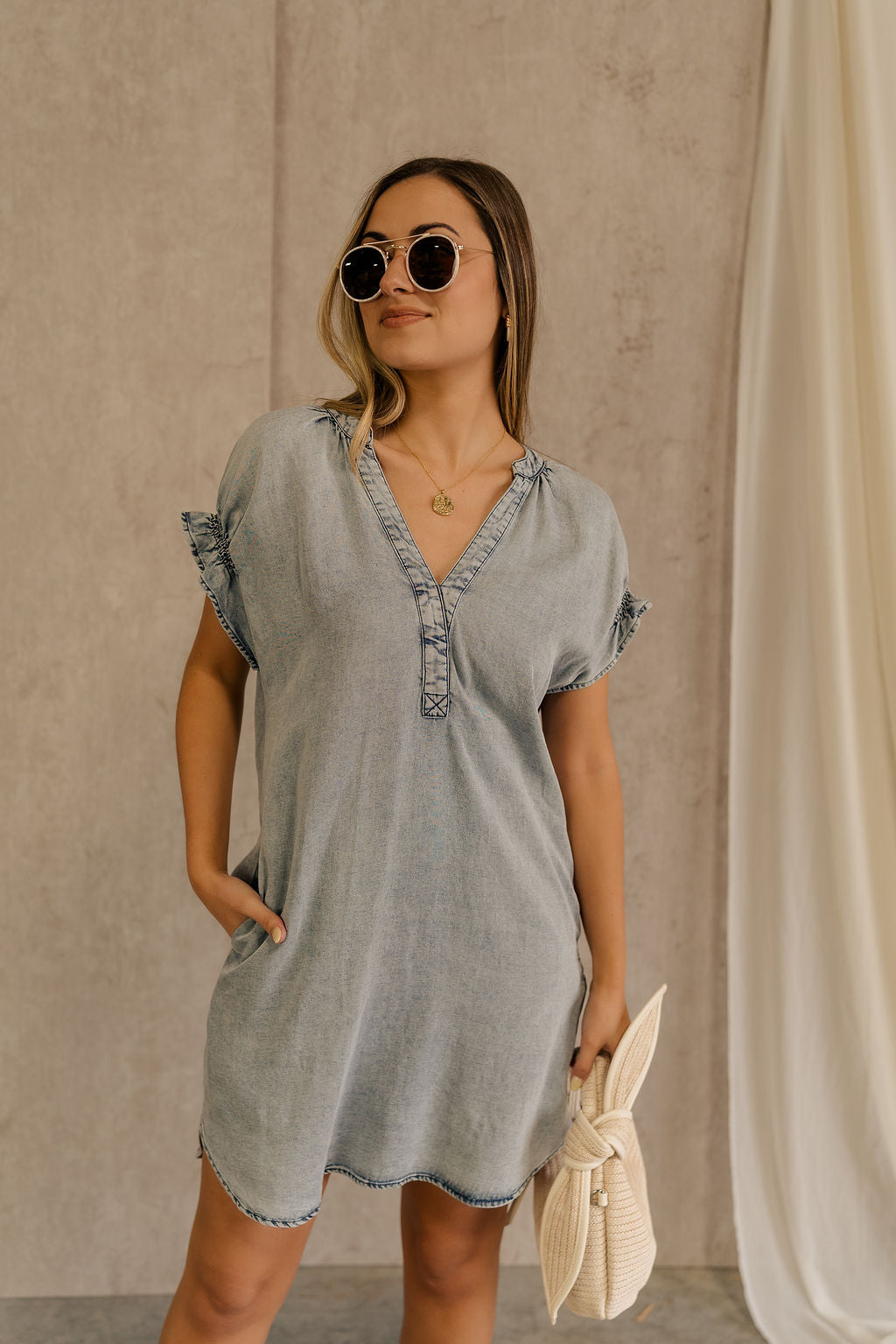 Front view of female model wearing the Leah Denim Wash Tencel Mini Dress which features denim blue tencel fabric, mini length, two side slit pockets, a round neckline with a v-cutout, an elastic band in the back, and short sleeves with elastic hems and ruffle details.