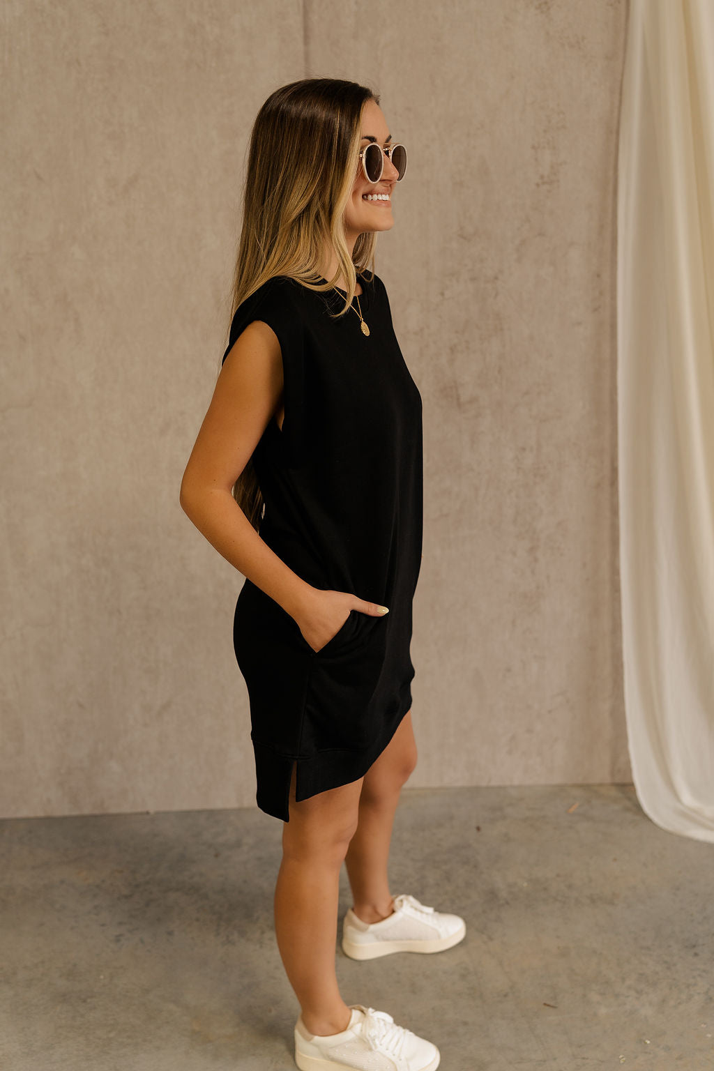 Full body view of female model wearing the Rayven Black Sleeveless Mini Dress which features black cotton fabric, mini length, ribbed hem, small side slits, two side slit pockets, a round neckline, and sleeveless.