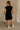 Full body back view of female model wearing the Rayven Black Sleeveless Mini Dress which features black cotton fabric, mini length, ribbed hem, small side slits, two side slit pockets, a round neckline, and sleeveless.