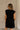 Back view of female model wearing the Rayven Black Sleeveless Mini Dress which features black cotton fabric, mini length, ribbed hem, small side slits, two side slit pockets, a round neckline, and sleeveless.