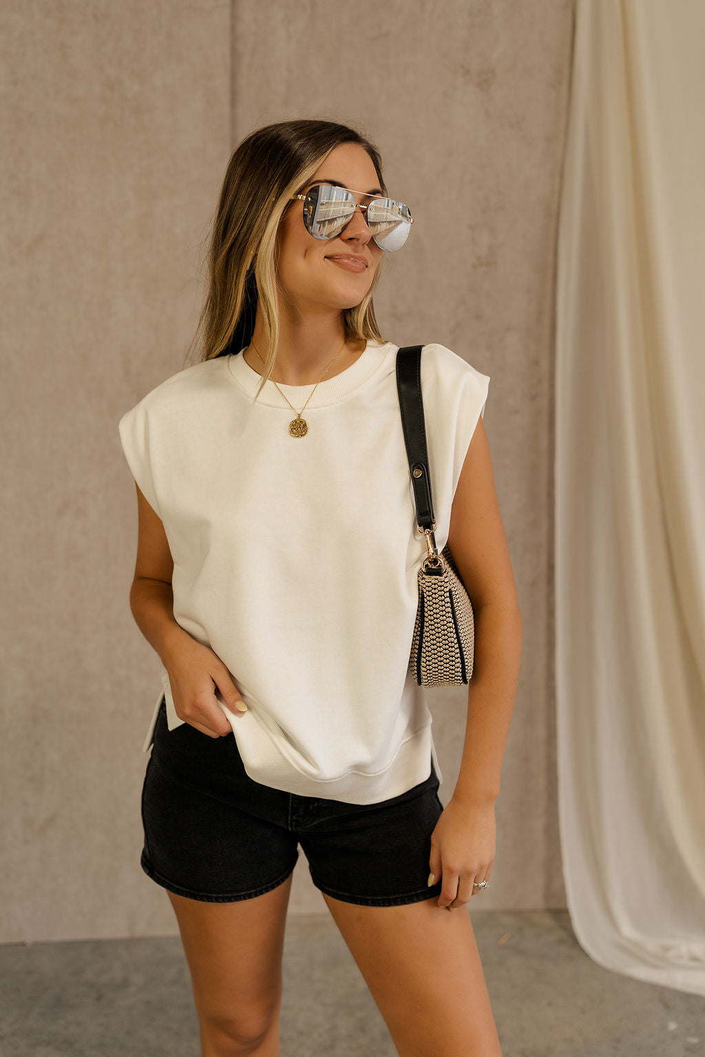 Front view of female model wearing the Ellie Off White Sleeveless Top which features white cotton fabric, ribbed hem, small side slits, round neckline and sleeveless