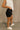 Side view of female model wearing the Ava Washed Black Denim Shorts which features Washed Black Denim Fabric, Two Front Pockets, Two Back Pockets, Silver Button with Zipper Closure and Belt Loops