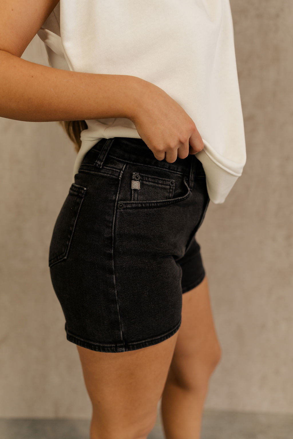 Side view of female model wearing the Ava Washed Black Denim Shorts which features Washed Black Denim Fabric, Two Front Pockets, Two Back Pockets, Silver Button with Zipper Closure and Belt Loops