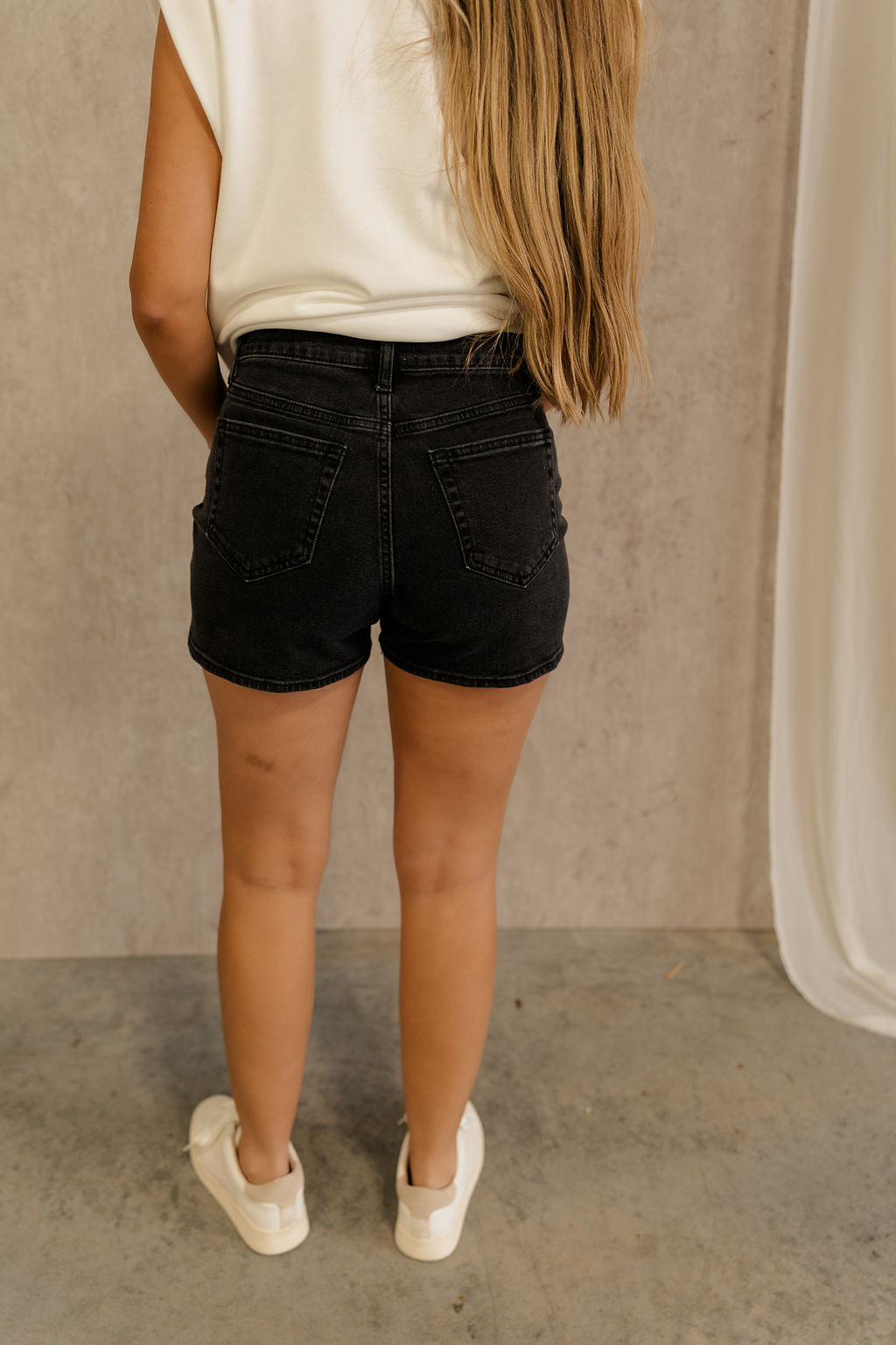 Back view of female model wearing the Ava Washed Black Denim Shorts which features Washed Black Denim Fabric, Two Front Pockets, Two Back Pockets, Silver Button with Zipper Closure and Belt Loops