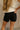 Back view of female model wearing the Ava Washed Black Denim Shorts which features Washed Black Denim Fabric, Two Front Pockets, Two Back Pockets, Silver Button with Zipper Closure and Belt Loops