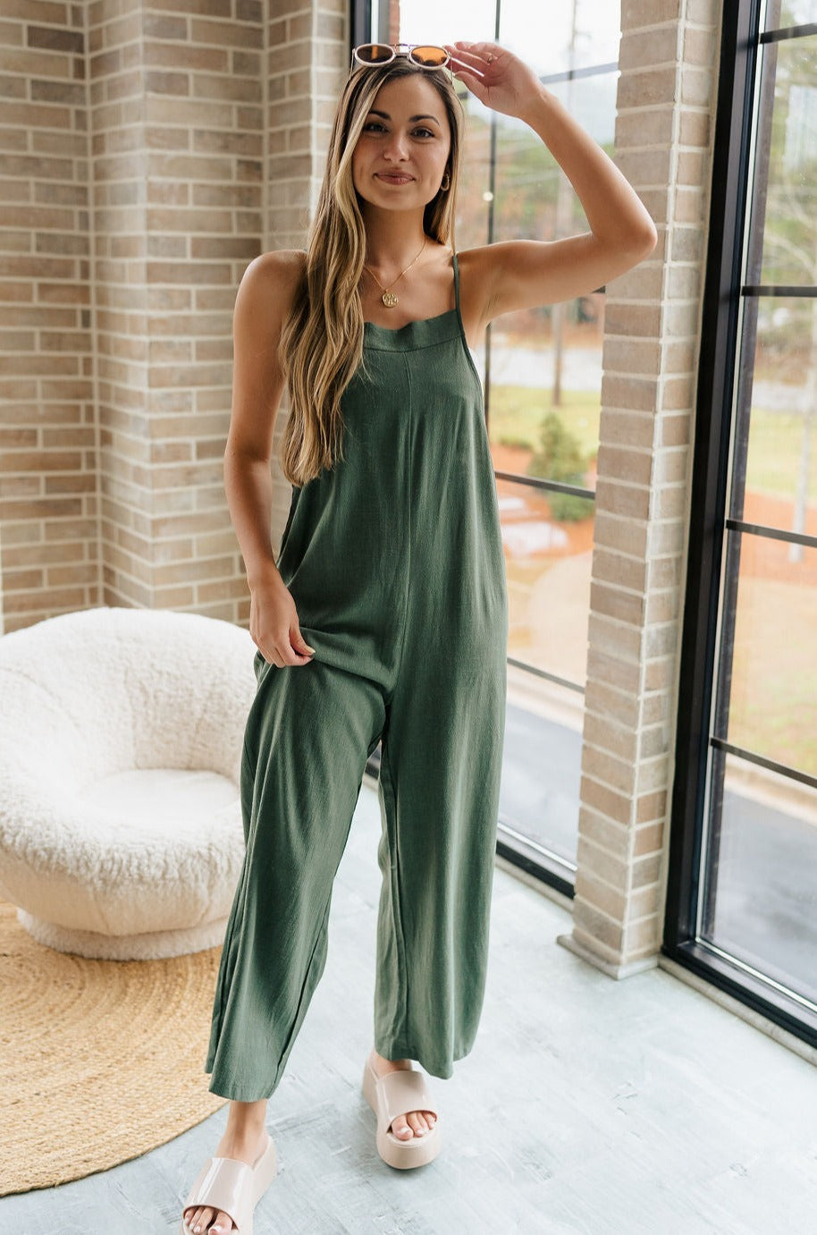 Full body front view of female model wearing the Jessie Wide Leg Jumpsuit in Green, that features dark green woven material, adjustable straps, and wide legs. Worn over white bandeau.