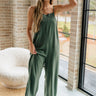 Full body front view of female model wearing the Jessie Wide Leg Jumpsuit in Green, that features dark green woven material, adjustable straps, and wide legs. Worn over white bandeau.