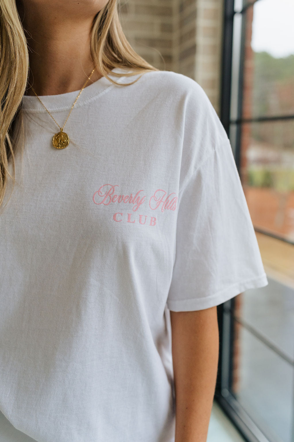 Close up view of female model wearing the Beverly Hills Tennis Club Graphic Tee which features White Cotton Fabric, Round Neckline , Short Sleeves, Beverly Hills Tennis Club in pink and green writing.