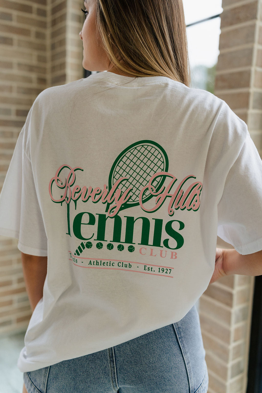 Back view of female model wearing the Beverly Hills Tennis Club Graphic Tee which features  White Cotton Fabric, Round Neckline , Short Sleeves, Beverly Hills Tennis Club in pink and green writing.