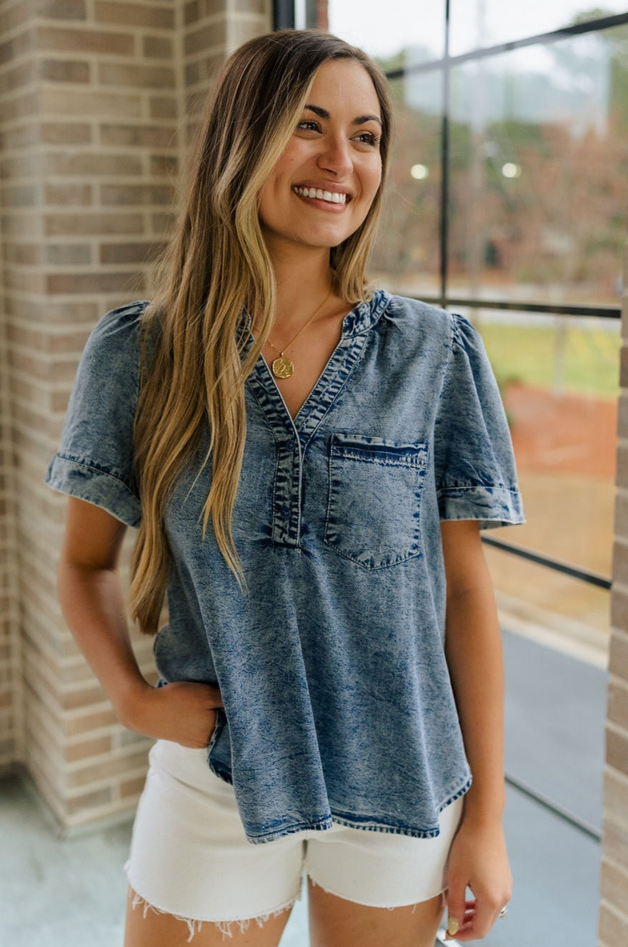 Front view of female model wearing the Tarah Medium Wash Short Sleeve Top which features washed denim tencel fabric, scooped hem, front left chest pocket, monochrome quarter button up, v-neckline and short sleeves