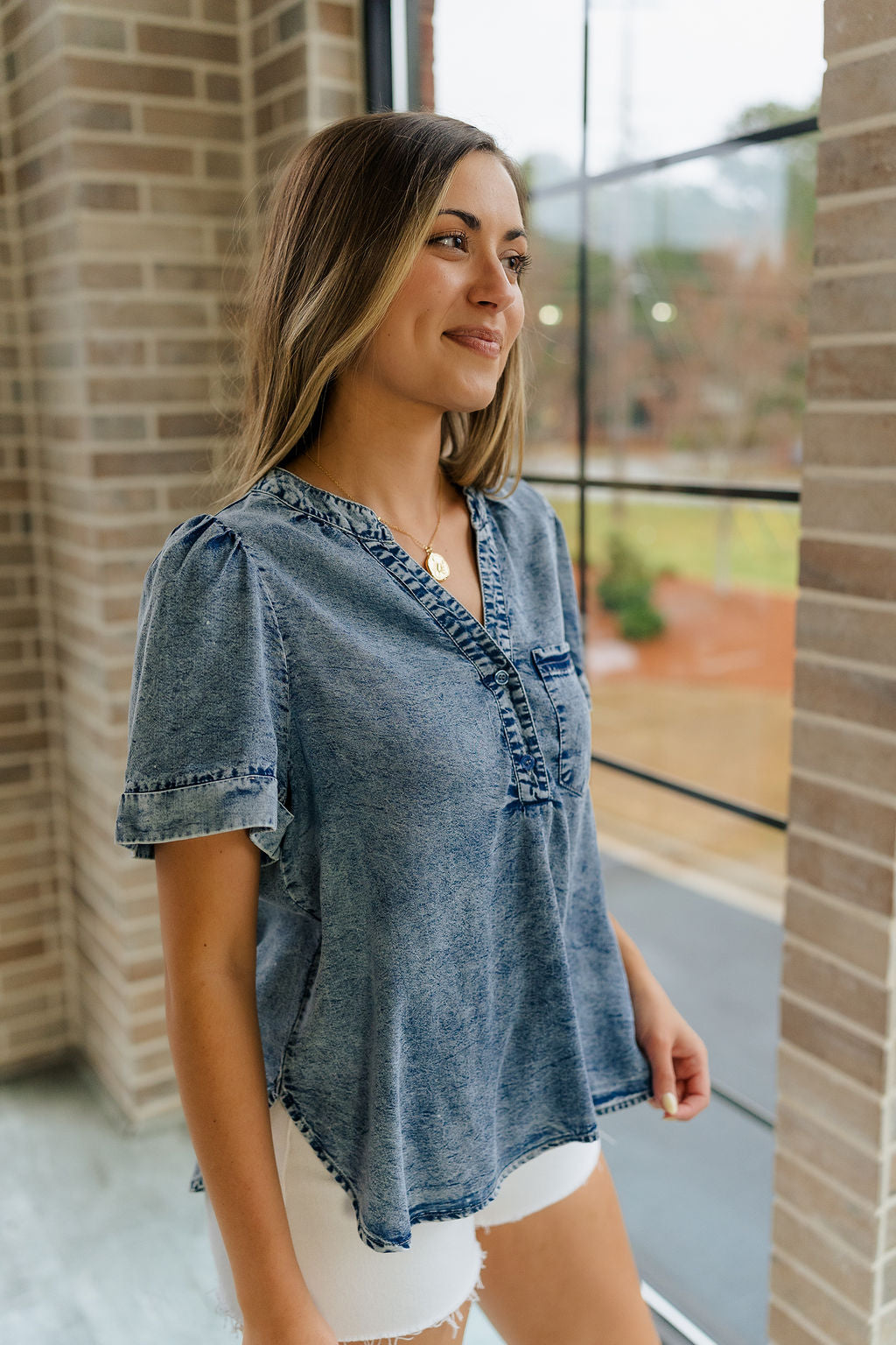 Side view of female model wearing the Tarah Medium Wash Short Sleeve Top which features washed denim tencel fabric, scooped hem, front left chest pocket, monochrome quarter button up, v-neckline and short sleeves