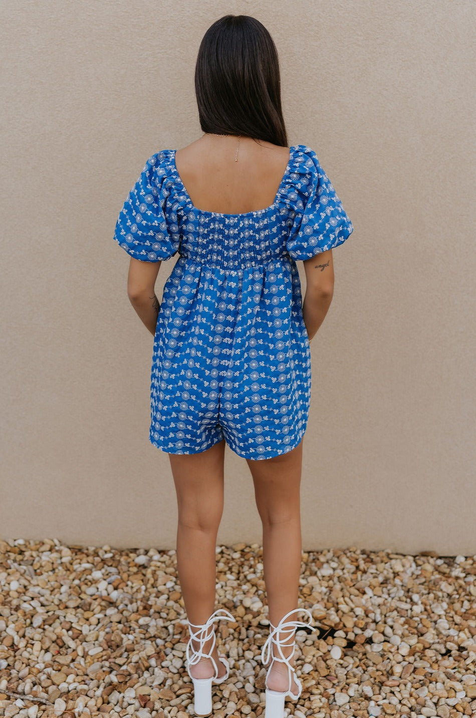 Full body back view of model wearing the Marissa Blue and White Floral Short Sleeve Romper which features Blue Lightweight Fabric, White Floral Design, Blue Lining, Square Neckline, Short Puff Sleeves and Smocked Back.