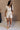 Full body view of female model wearing the Kristin Oatmeal Linen Short Sleeve Romper which features Oatmeal Linen Fabric, Two Front Pockets, Front Snap Buttons, Collared Neckline and Short Puff Sleeves.