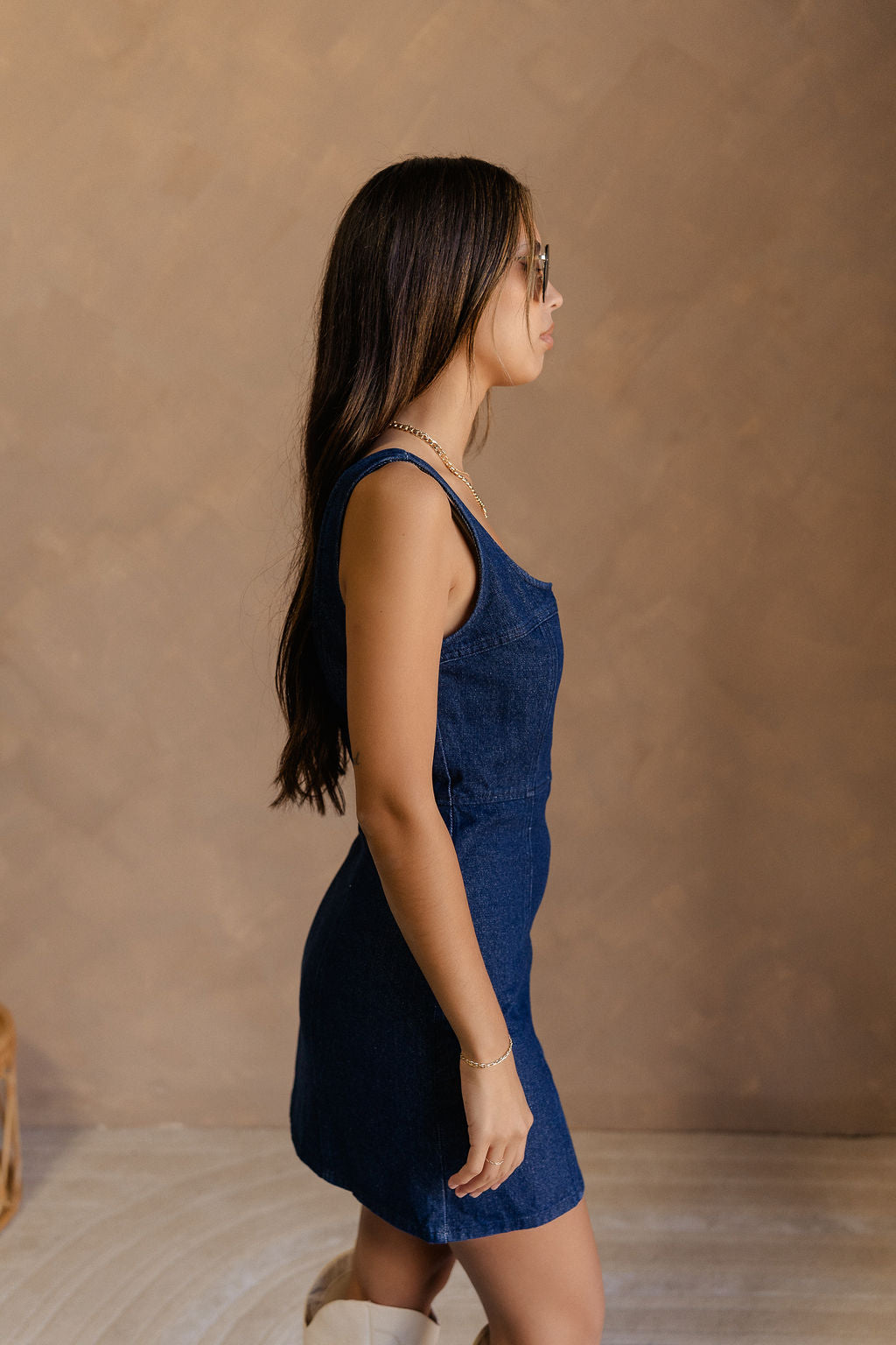 Side view of female model wearing the Stella Dark Denim Wash Sleeveless Mini Dress which features features Dark Denim Wash Fabric, Front Zip-Up Closure, Square Neckline and Sleeveless.