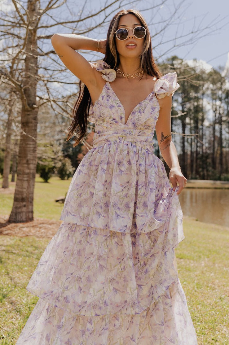 front view of model wearing The Harlow Lavender Floral Ruffle Midi Dress features sheer fabric with purple, green, orange floral print, shimmer thread details, beige lining, a tiered body with a maxi-length hem, ruffled straps, a plunge neckline, a lace-up back, and a back zipper and hook closure.