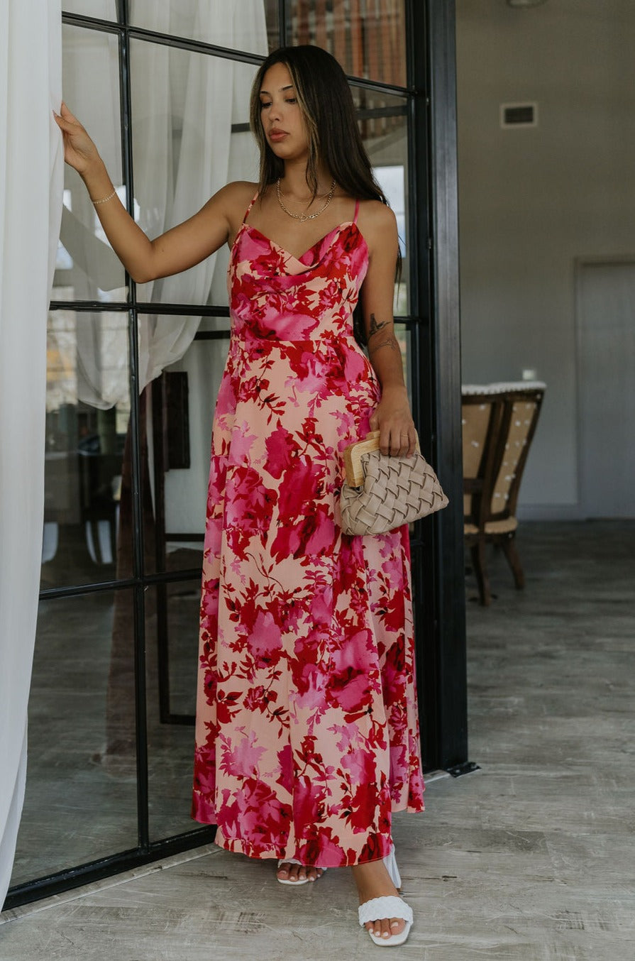 Full body front view of model wearing the Roselyn Pink & Red Floral Maxi Dress that has light pink fabric with dark pink and red floral pattern, a cowl neckline, and spaghetti straps that cross in the back.
