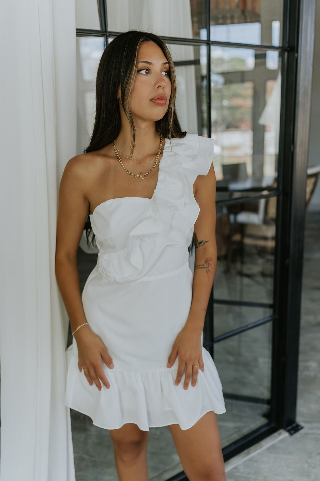 Front view of model wearing the Elyse White Ruffled One Shoulder Dress that has white fabric, one shoulder straps with ruffles, and a ruffled mini length hem.