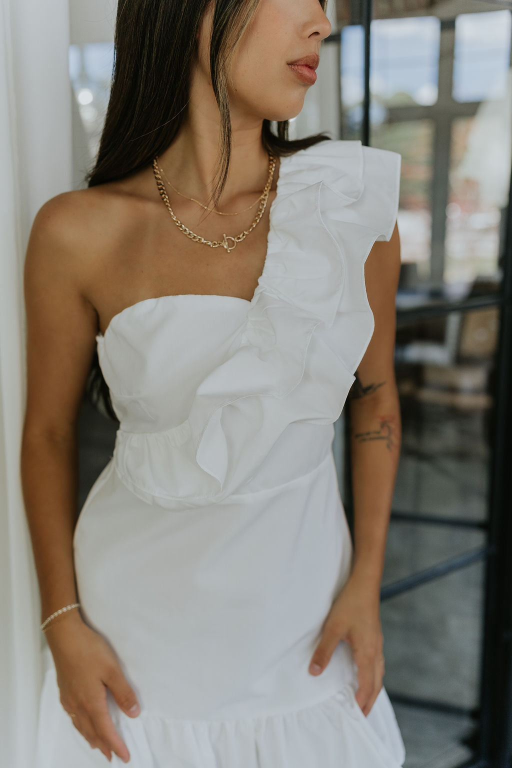 Close upper body front view of model wearing the Elyse White Ruffled One Shoulder Dress that has white fabric, one shoulder straps with ruffles, and a ruffled mini length hem.