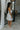 Full body right side view of model wearing the Elyse White Ruffled One Shoulder Dress that has white fabric, one shoulder straps with ruffles, and a ruffled mini length hem.