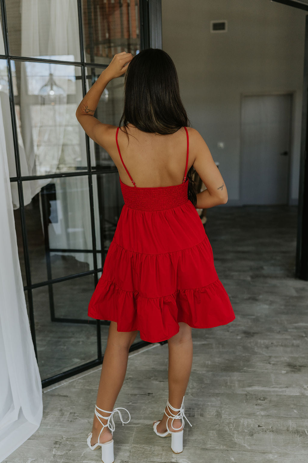 full body back view of model wearing the Loretta Red Sleeveless Mini Dress that has a tiered skirt, surplice neckline, and thin straps. Worn with white heels.
