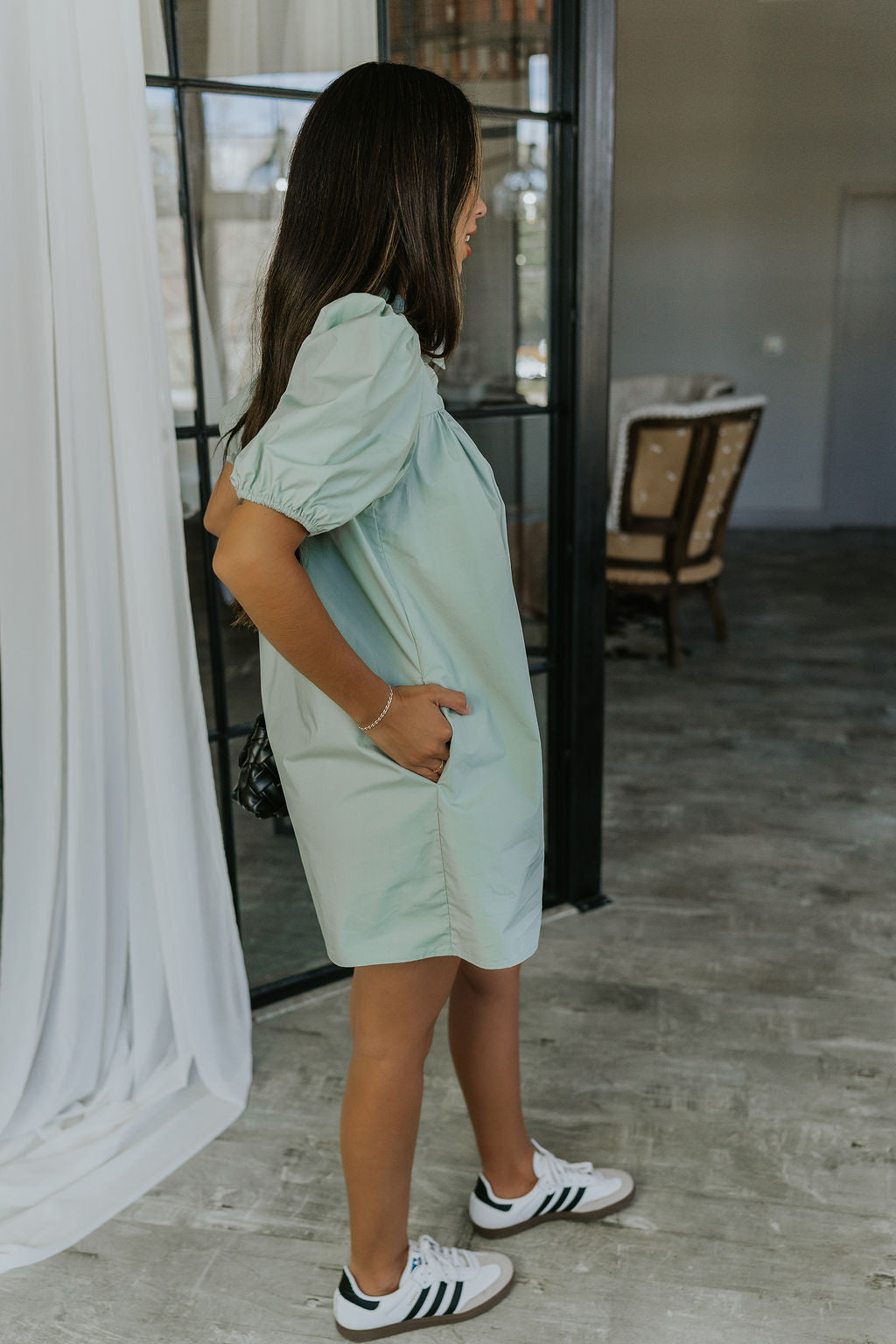 Full body side view of model wearing the Marianna Sage Short Sleeve Dress that has sage green fabric, a button-up front, collar, and short puff sleeves.