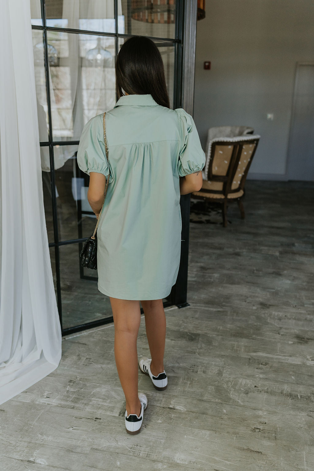 Full body back view of model wearing the Marianna Sage Short Sleeve Dress that has sage green fabric, a button-up front, collar, and short puff sleeves.