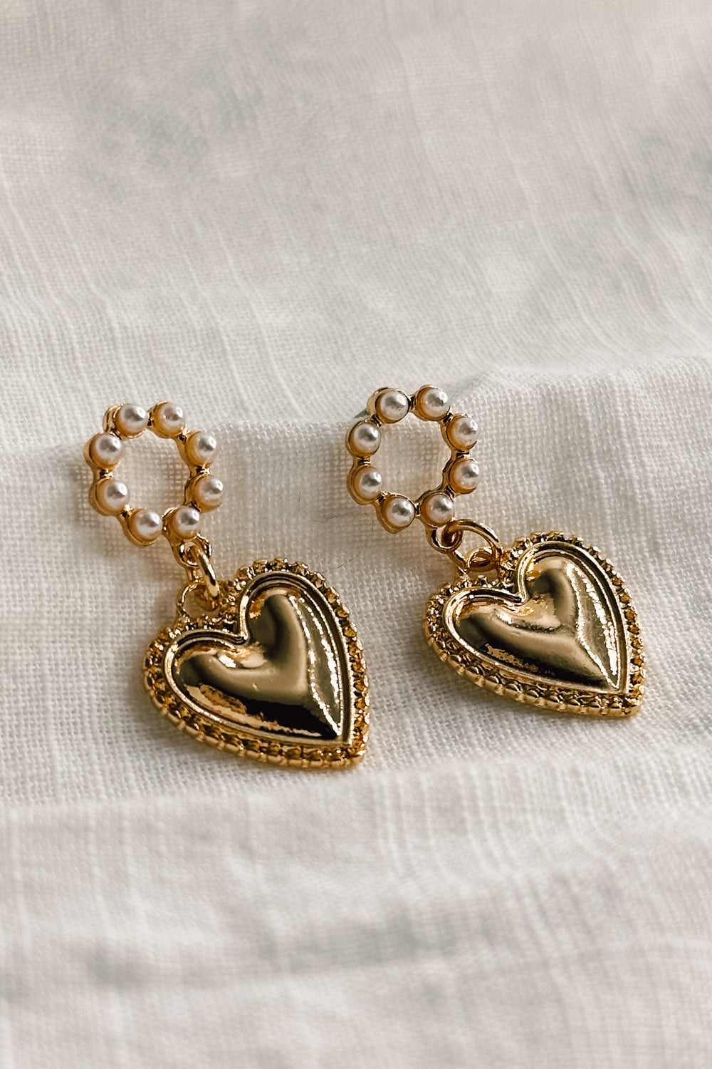Close up view of the Brielle Pearl & Gold Heart Shaped Dangle Earring which features gold heart shaped dangle medallion and pearl details