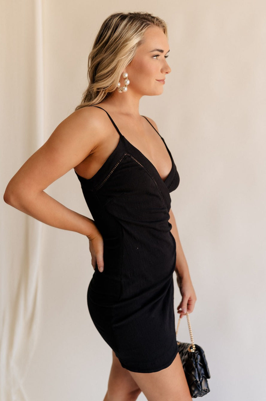 Side view of female model wearing the Sophia Black Plunge Neckline Mini Dress which features Black Lightweight Fabric, Mini Length, Side Ruched Detail with Covered Buttons Closure, Scoop Neckline, Adjustable Straps and Sleeveless