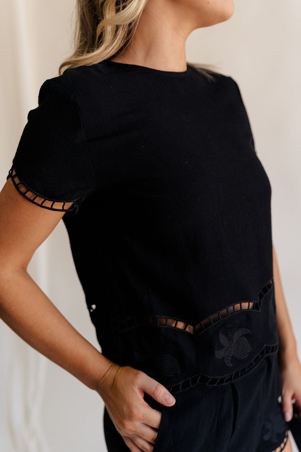 Close up side view of female model wearing the Stella Black Floral Trim Short Sleeve Top which features Black Knit Fabric, Cropped Waist, Scalloped Hem with Open Details, Monochrome Floral Print, Short Sleeves, Round Neckline and Back Button Closure