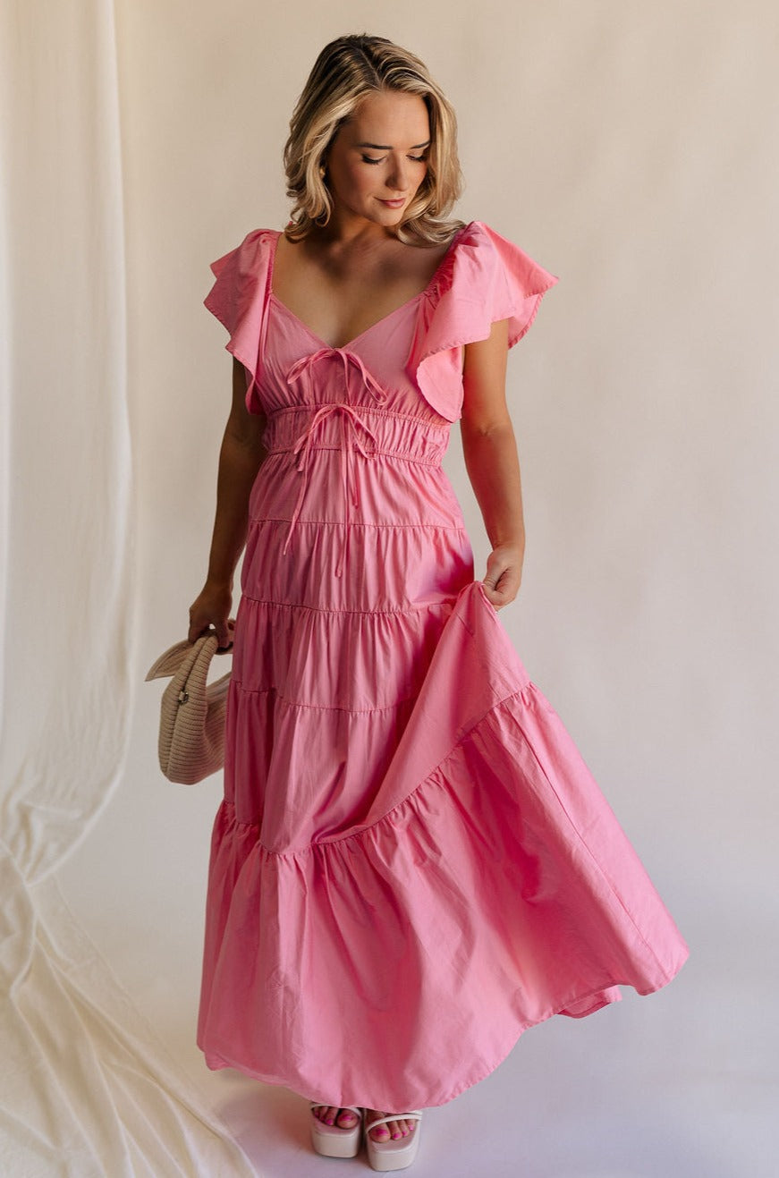 Full body view of female model wearing the Gianna Ruffle Short Sleeve Tiered Maxi Dress in PInk which features Lightweight Fabric, Tiered Body, Midi Length, Pockets on each side, V-Neckline with a Tie Detail and Ruffle Short Sleeves. the dress is available in pink, aqua blue and white.