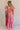 Full body back view of female model wearing the Gianna Ruffle Short Sleeve Tiered Maxi Dress in PInk which features Lightweight Fabric, Tiered Body, Midi Length, Pockets on each side, V-Neckline with a Tie Detail and Ruffle Short Sleeves. the dress is available in pink, aqua blue and white.