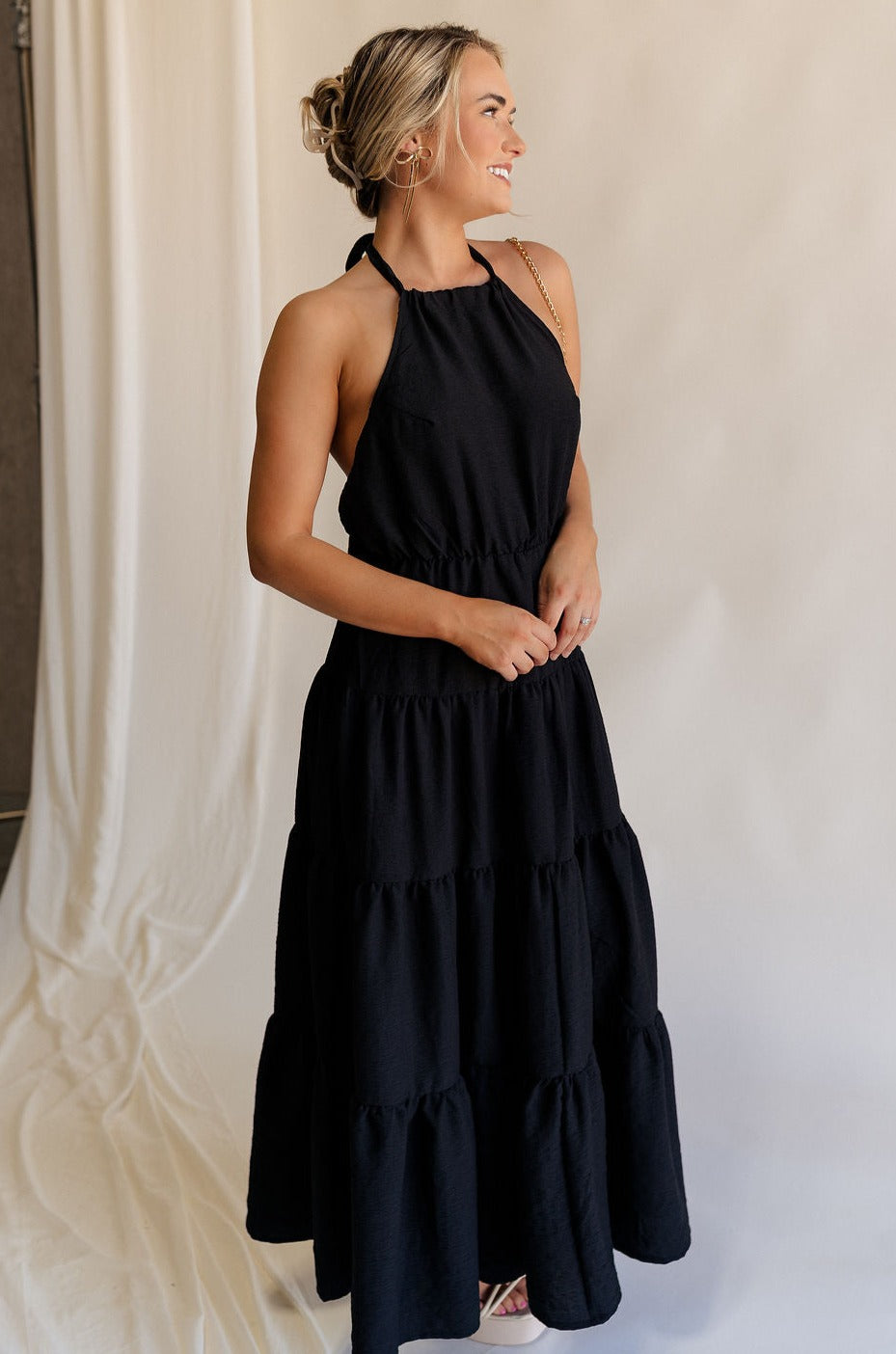 Full body side view of female model wearing the Luna Halter Tie Tiered Maxi Dress in black which features Lightweight Fabric, Tiered Body, Maxi Length, Thigh Length Lining, Halter Neckline with Tie and Open Back