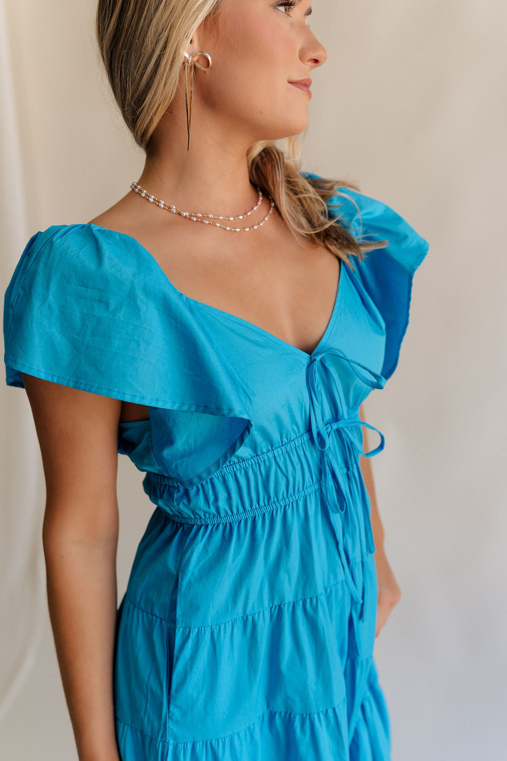 Close up side view of female model wearing the Gianna Ruffle Short Sleeve Tiered Maxi Dress in Blue which features Lightweight Fabric, Tiered Body, Midi Length, Pockets on each side, V-Neckline with a Tie Detail and Ruffle Short Sleeves. the dress is available in pink, aqua blue and white.