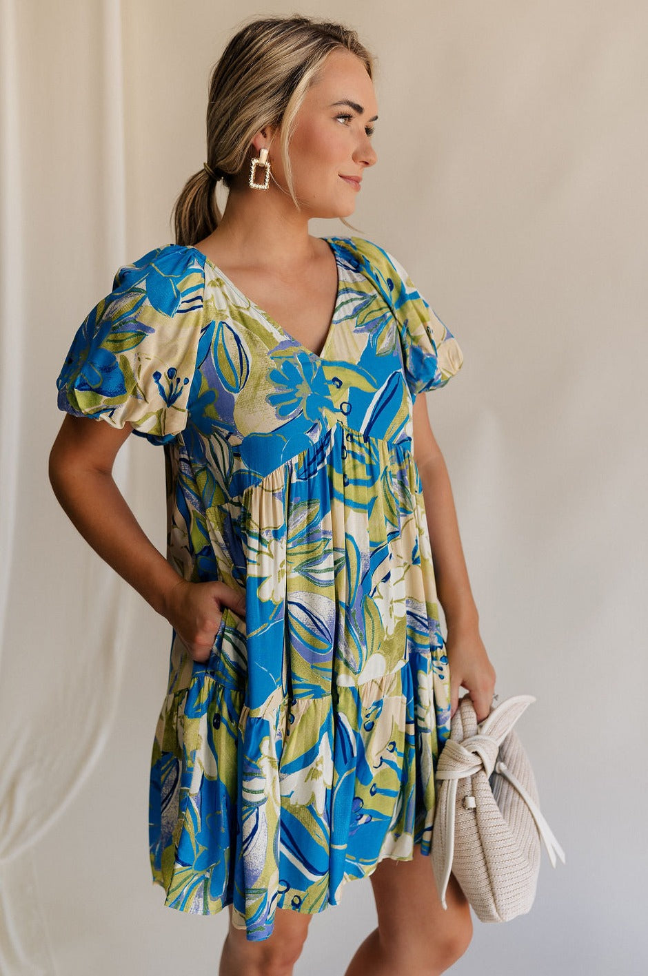 Side view of female model wearing the Tessa Blue & Lime Green Floral Mini Dress which features Blue, Green and White Light Weight Fabric, Floral Print, Mini Length, Tiered Body, Blue Lining, V-Neckline and Short Puff Sleeves