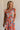 Front view of female model wearing the Kai Orange Multi Floral Midi Ruffle Dress which features Orange, Blue, Green, Purple and Yellow Lightweight Fabric, Floral Design, Tiered Body, Orange Lining, Midi Length, Pockets On Each Side, V-Neckline and Ruffle Short Sleeves