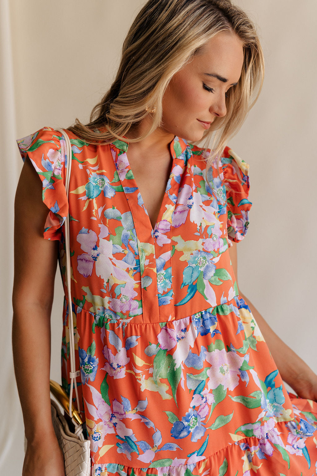 Close up view of female model wearing the Kai Orange Multi Floral Midi Ruffle Dress which features Orange, Blue, Green, Purple and Yellow Lightweight Fabric, Floral Design, Tiered Body, Orange Lining, Midi Length, Pockets On Each Side, V-Neckline and Ruffle Short Sleeves