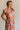 Side view of female model wearing the Kai Orange Multi Floral Midi Ruffle Dress which features Orange, Blue, Green, Purple and Yellow Lightweight Fabric, Floral Design, Tiered Body, Orange Lining, Midi Length, Pockets On Each Side, V-Neckline and Ruffle Short Sleeves