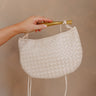 Hand is holding the Nadia Woven Purse on her shoulder. It features a cream woven body topped with a gold bar accent and cream straps.