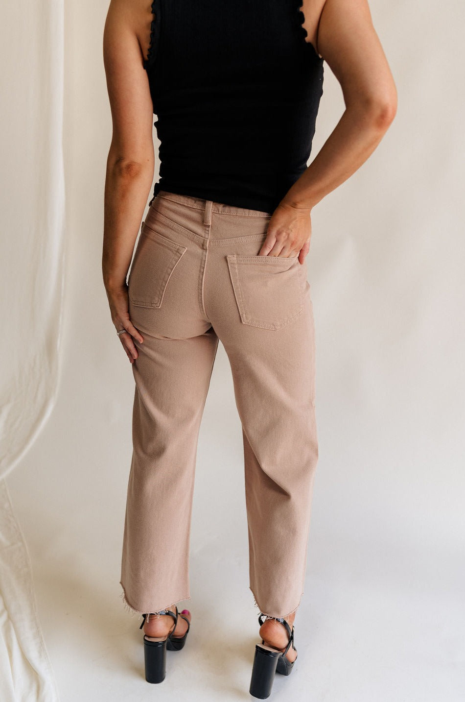 Back view of female model wearing the Lena Light Taupe Wide Cropped Leg Pants which features Light Taupe Denim Fabric, Wide Pant Leg, Raw Hem, Front Zipper with Button Closure, Two Front Pockets, Two Back Pockets and Belt Loops