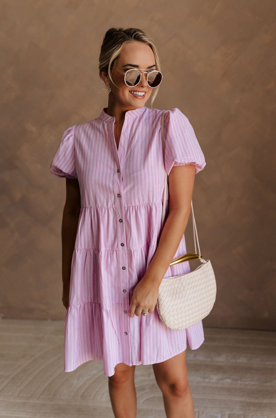 Upper body front view of female model wearing the Cameron Pink & White Striped Dress that has pink and white vertical striped fabric, short puff sleeves, a tiered skirt, and button up front.