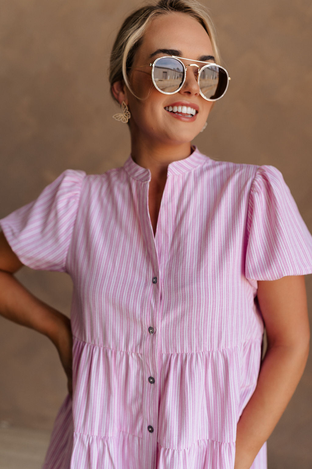 Upper body front view of female model wearing the Cameron Pink & White Striped Dress that has pink and white vertical striped fabric, short puff sleeves, a tiered skirt, and button up front.