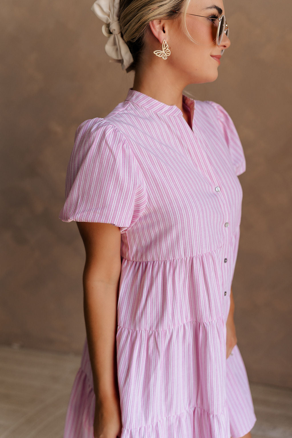 Upper body side view of female model wearing the Cameron Pink & White Striped Dress that has pink and white vertical striped fabric, short puff sleeves, a tiered skirt, and button up front.