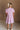 Full body back view of female model wearing the Cameron Pink & White Striped Dress that has pink and white vertical striped fabric, short puff sleeves, a tiered skirt, and button up front.