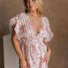 Upper body front view of female model wearing the Leona Pink Floral Romper that has pink sheer fabric with a floral pattern, pink lining, plunge neck, and short ruffled sleeves.