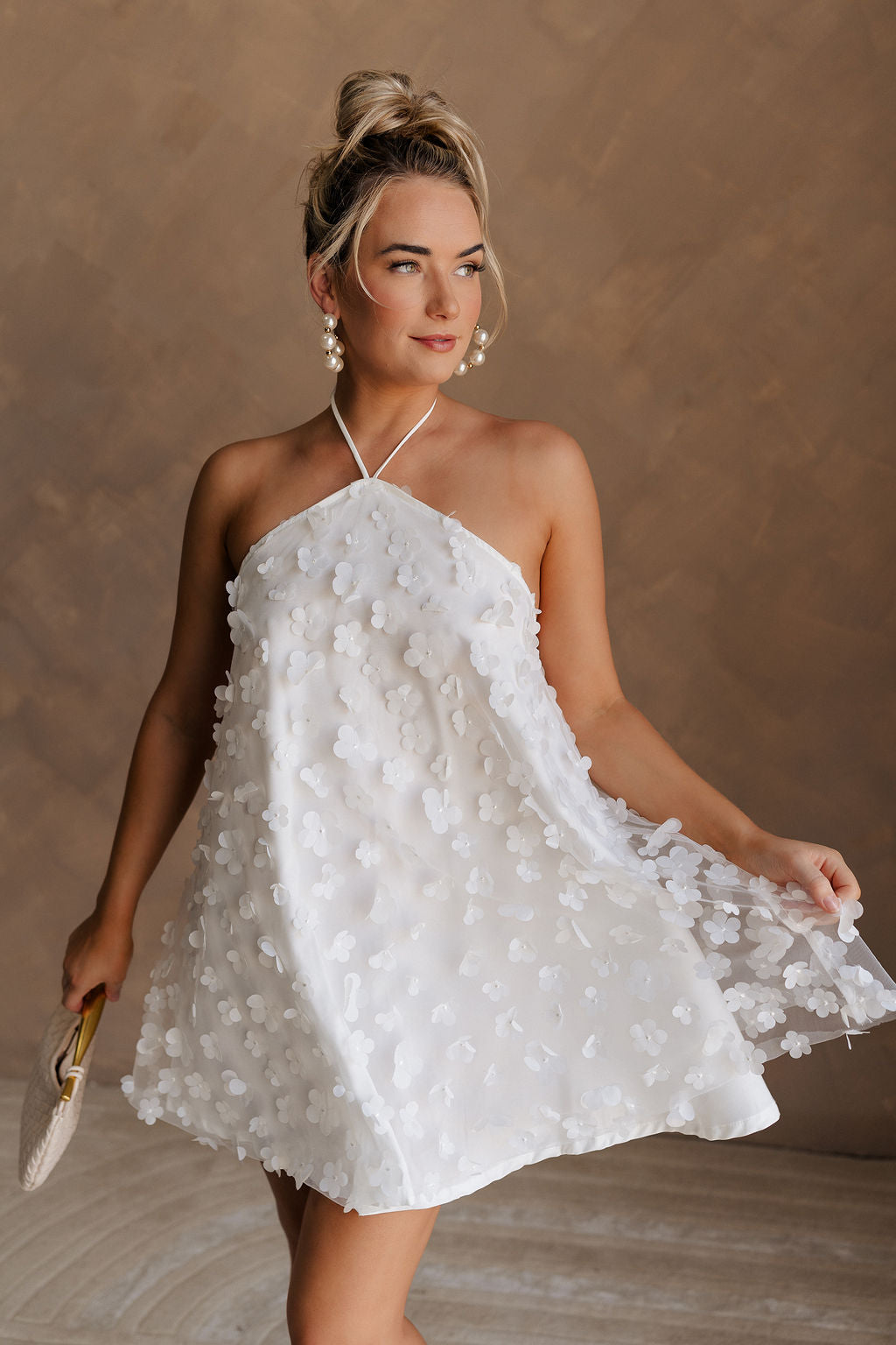 Upper body front view of female model wearing the Meredith White Embellished Floral Dress that has white sheer fabric with 3d floral embellishments, white lining, and a halter neck.