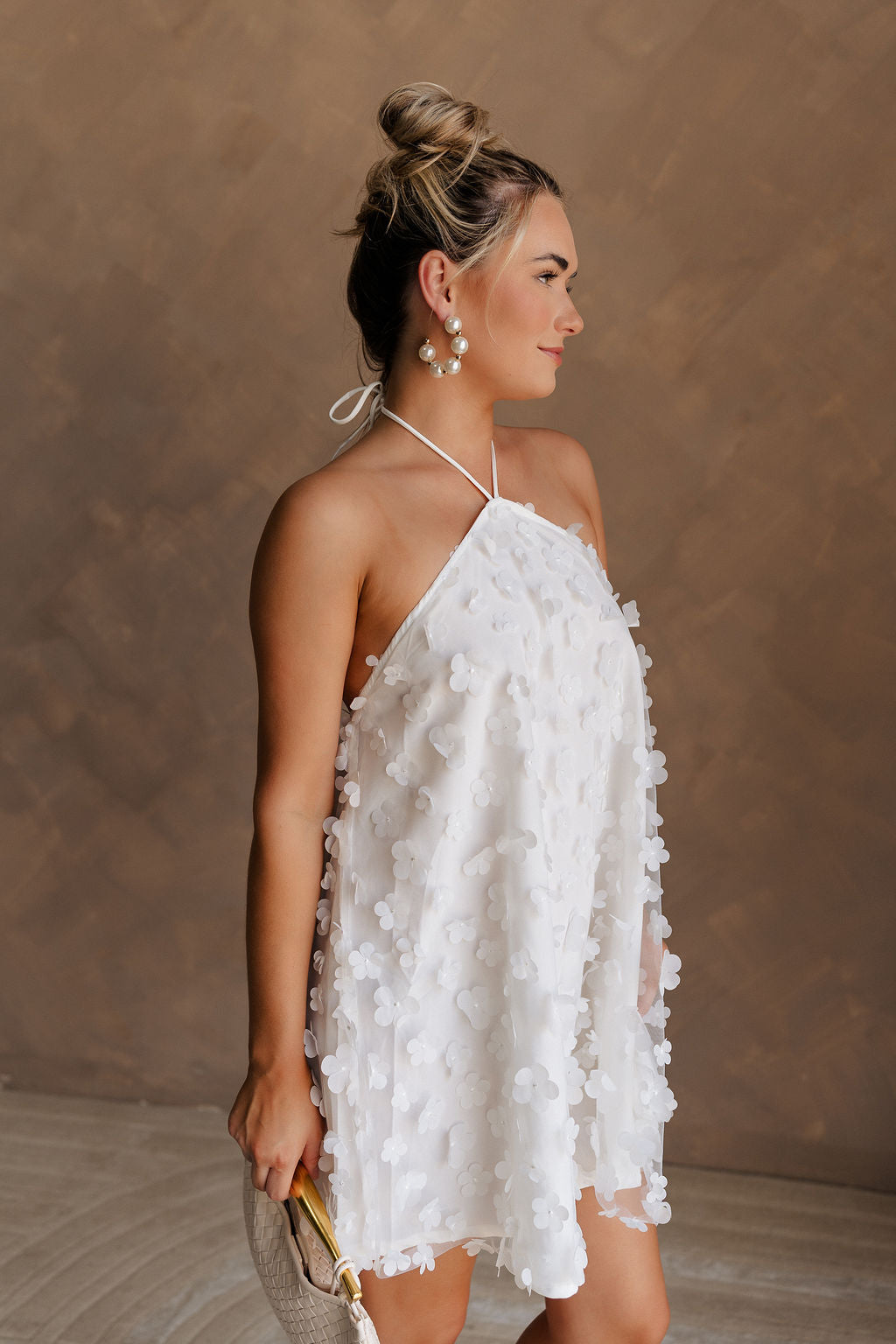 Upper body side view of female model wearing the Meredith White Embellished Floral Dress that has white sheer fabric with 3d floral embellishments, white lining, and a halter neck.