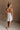 Full body back view of female model wearing the Meredith White Embellished Floral Dress that has white sheer fabric with 3d floral embellishments, white lining, and a halter neck.